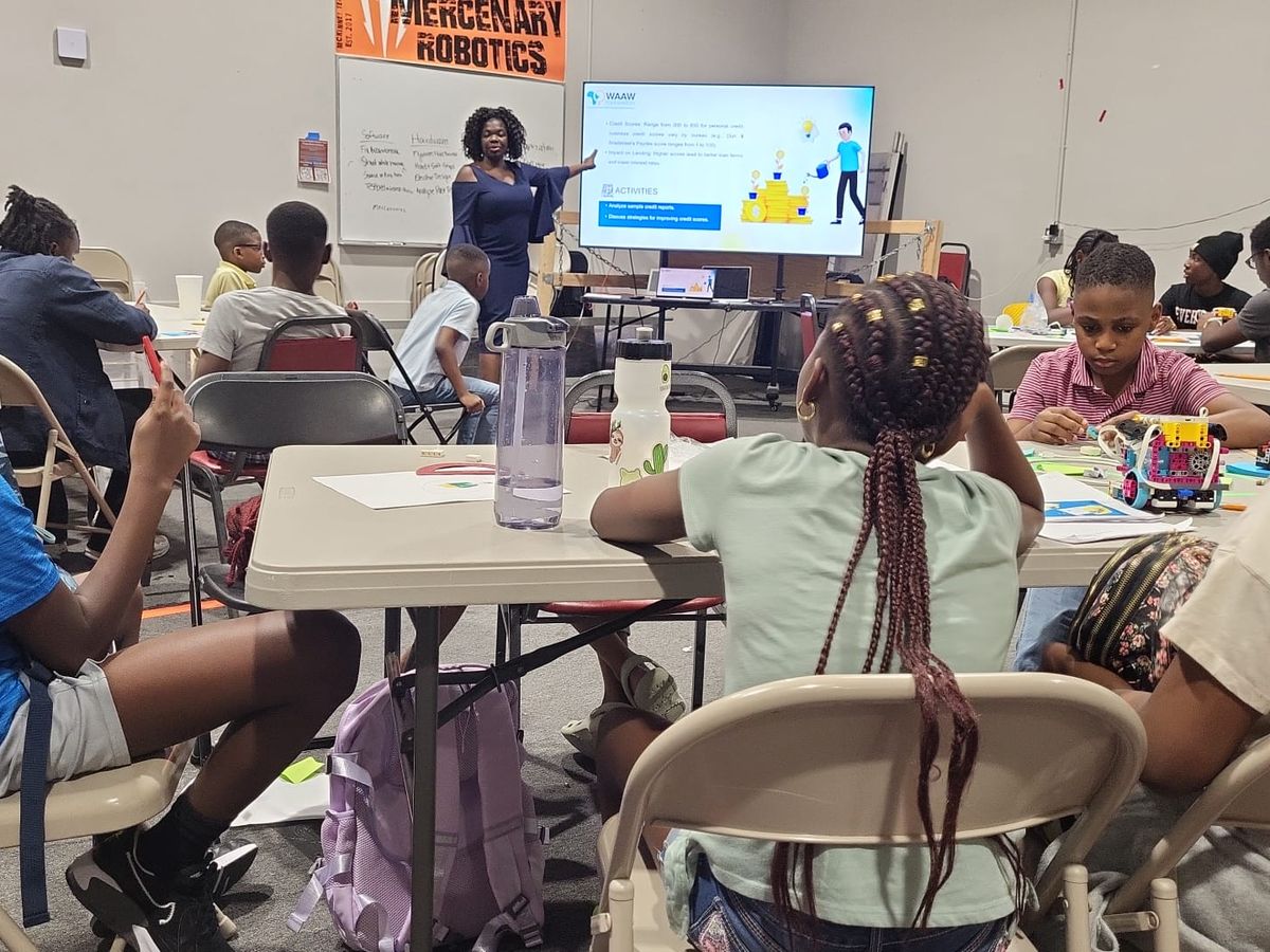 Kids at tables with their robots while a presenter teaches at a screen